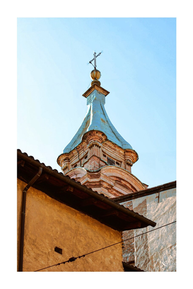 The image depicts the top of a church tower against a pale blue sky. The tower is ornate, with a light blue, conical roof and a cross at the very top. Parts of buildings with walls and roofs are visible in the foreground.