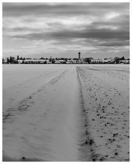 Blick über ein verschneites Feld zu einer kleinen Stadt mit dunklem Himmel.
