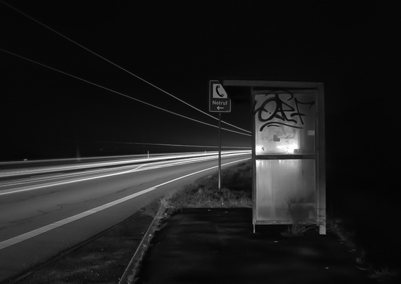 Man sieht eine Bushaltestelle auf der rechten Seite. Links davon, an der Bundesstrasse, sieht man bedingt durch eine Langzeitbelichtung, nur noch weiße Stiche der vorüber fahrenden Autos und LKW´s. Das Ganze ist in der Nacht aufgenommen worden und das Foto ist in Schwarzweiss.