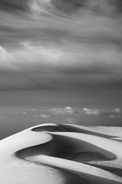 vertical black and white dune shot. Dune with multiple curves and curved shadows at the bottom, dramatic sky at the top.