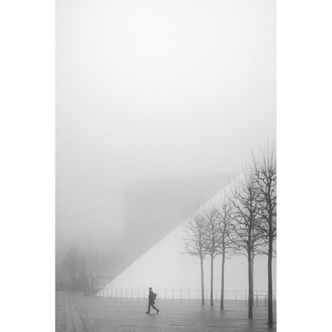 Black and white street photography taken in Luxembourg. The silhouette of a man walking towards the congress center appears in a foggy background composed of the back of the Philharmonie and a few naked trees.