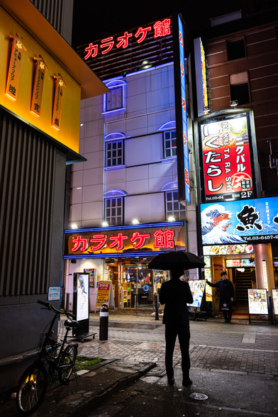 A quiet street bathed in the red and purple neon lights of a karaoke place in Shimbashi, in Tokyo, at night. In the foreground the silhouette of a man in a dark costume standing and holding a black umbrella. Opposite him next to the Karaoke, a restaurant waiter, also dressed in black, is taking a break in front of his shop.