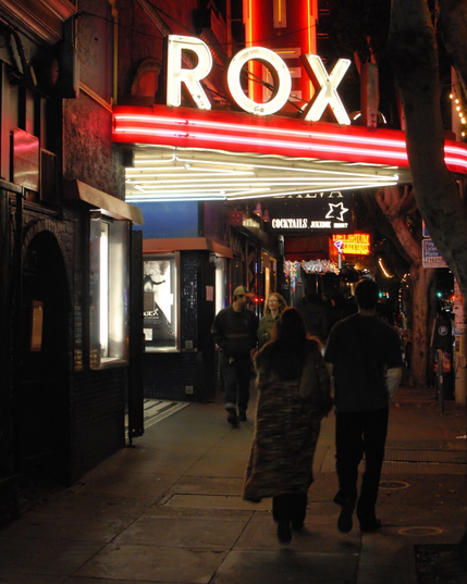 A night photo outside the Roxie Theater. The lighting is dominated by the neon Roxie sign. Young couples walk past on their way to the next bar