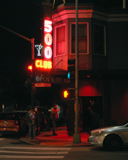 Two men chat animatedly outside a club, gesticulating, while another man stands off to the side smoking. The scene is dark, lit only by the club's neon sign and a traffic light