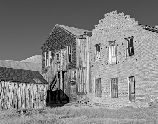 A black and white landscape photo of the backs of two old abandoned buildings. Both are two stories. The one on the right is made of brick while the one on the left is made of wood. The one of the right has two windows with a door in the middle on the second floor, but the porch is missing. The ground floor has two windows on the left with a door on the right. The wood building has two  windows on the upper floor with a enclosed staircase leading from a central door down to the left disappearing behind a wood shed on the far left. The sky is very dark simulating the use of a red filter in black and white photography. Long dry grass is in the foreground.