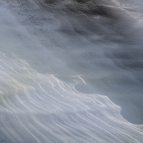 Das Foto zeigt eine von der Sonne beleuchtete, eisbedeckte Landschaft. Die Eisfläche ist nicht gleichmäßig, sondern weist sanfte Dünen auf, die durch den Wind geformt wurden. Licht und Schatten spielen auf der Oberfläche und erzeugen ein fast abstraktes Muster aus hellen und dunklen Linien. Im Hintergrund ist eine Wasserfläche auf dem Eis zu sehen, die durch den Wind gewelllt wird.  Das Bild wirkt sehr ruhig und beinahe surreal.
 -------- 
The photo shows a sunlit, ice-covered landscape. The ice surface is not uniform, but features gentle dunes formed by the wind. Light and shadow play on the surface, creating an almost abstract pattern of light and dark lines. In the background, a body of water can be seen on the ice, rippled by the wind.  The image appears very calm and almost surreal.