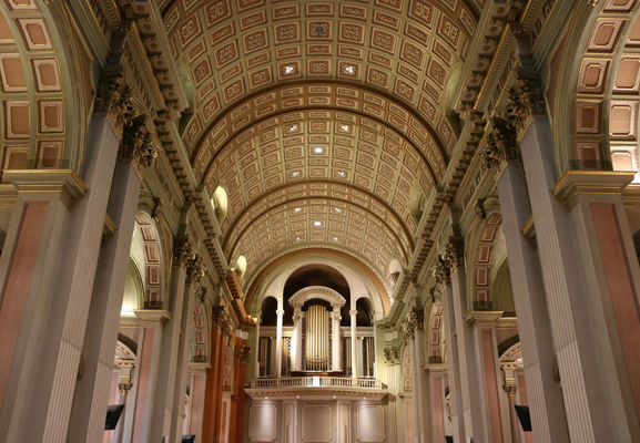 Photograph of the central aisle of a cathedral, showing its arched ceiling adorned with square and rectangular panels decorated with gilded moldings, square columns with Corinthian capitals on either side, and its majestic organ on its mezzanine at the far end. Openings to the side aisles are visible. The pink, brown, pale green, and gold create a soft effect.

Photographie de l'allée centrale d'une cathédrale, montrant son plafond en arche orné de planneaux carrés et rectanguaires ornés de décorations et moulures dorées, des colonnes carrées avec des chapiteaux corinthiens de chaque côté, et son majestueux orgue sur sa mezzanine tout au fond. Des ouvertures vers les ailes secondaires sont visibles. Le rose, le brun, le vert pâle et le doré ont un effet doux.