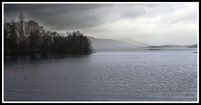 A serene lake scene under a cloudy sky, featuring dark water and silhouettes of trees along the shoreline. In the distance, hills are partially obscured by mist. An orange buoy floats in the foreground, while remnants of a dock are visible across the water.