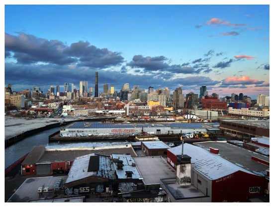 The skyline of downtown Brooklyn at sunset, as seen from the elevated platform of the Smith-9th Street station overlooking the Gowanus Canal. Low warehouses with graffiti scrawled on their walls occupy the foreground. At left is a glimpse of the canal curving around and in between the warehouses. In the distance is the city skyline, with the supertall Brooklyn Tower thrusting up far above the other buildings. A low mass of clouds — their edges illuminated by the setting sun behind camera — hovers over the buildings, eventually giving way to a clear blue sky.