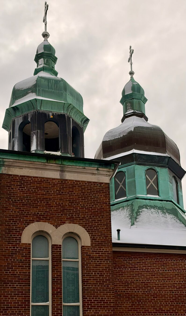 Two cupolas on top of a religious building; one has open arches, the other  is enclosed with windows. Both have a cross on top but the angle of the photo makes them visible only from an oblique. They have slightly ornate roofs and copper cladding. The building below is brown brick with two tall windows. Beige-coloured brick highlights the top of the windows in 2 arcs and along the straight roof line.  The sky is a dull grey, there is snow on the roof, and no other buildings are visible.