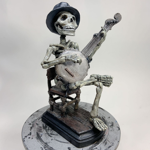 A painted polymer clay sculpture of a skeleton wearing a fedora sitting in a wooden chair and playing a old-style gourd banjo.