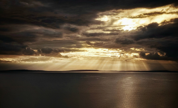 This photo is of Galway Bay. The sky is filled with dramatic, dark clouds, but the sun is setting, casting golden rays through the breaks in the cloud cover. These sunbeams stretch down and reflect on the water's surface. A distant landmass is visible on the horizon. (Galway and the Aran Islands)