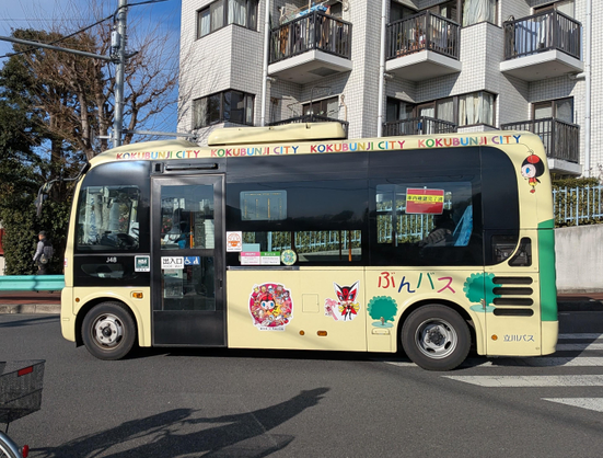 Gelber Kokubunji City Bus mit Maskottchen an der Seite. Passiert gerade eine Kreuzung in gleichnamiger Stadt in der Mitte der Präfektur Tokio.