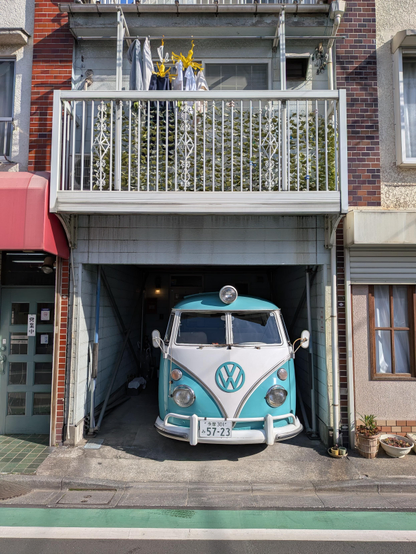 Ein blau-weisser VW Bulli Nachbau schaut mit der Front aus einer Garage heraus. Darüber der Balkon eines typischen kompakten Hauses in Japan. 