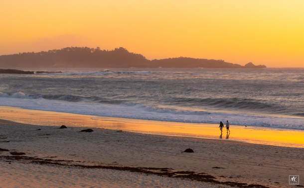 Color photo of two people walking along a beach that is reflecting  the yellow-orange colors of the sky above, lit by the setting sun.