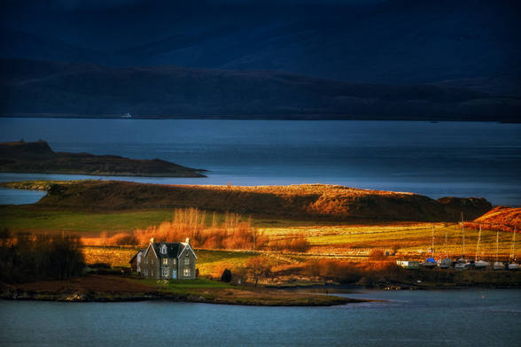 Ein traditionelles Steinhaus steht prominent am Ufer einer Bucht nahe Oban, Schottland. Die Szene wird von warmem, goldenem Licht beleuchtet, das die umliegenden Felder und Hügel hervorhebt, während der Hintergrund in tiefen Blautönen liegt. Boote sind am Ufer einer nahegelegenen Anlegestelle zu sehen, was die maritime Umgebung unterstreicht.
