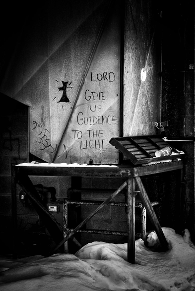 A message written on a wall saying "Lord give us guidance to the light" is seen along a snow covered alley. This is a black and white image.