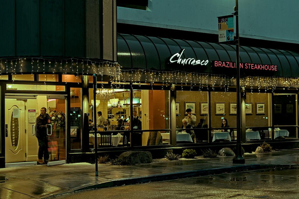 A color landscape photo of the front of a restaurant on a rainy night. A man is exiting the glass doors on the left. Just to the right of the doors you can see into the restaurant with waiters and people sitting at tables. To the left of the is street view seating behind a long series of glass windows. Most of the tables are empty except for one couple sitting at a table in the center. A waiter stands at the table taking orders or maybe serving wine. The sign over the outside awing reads, "Churrasco Brazillian Steakhouse." A small section of a wet reflective street is in the foreground.