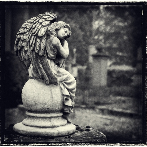 Ein schlafender Engel mit angelegten Flügeln auf einem Grabstein. Das Foto ist monochrome und schaut nach einer alten Fotografie aus.