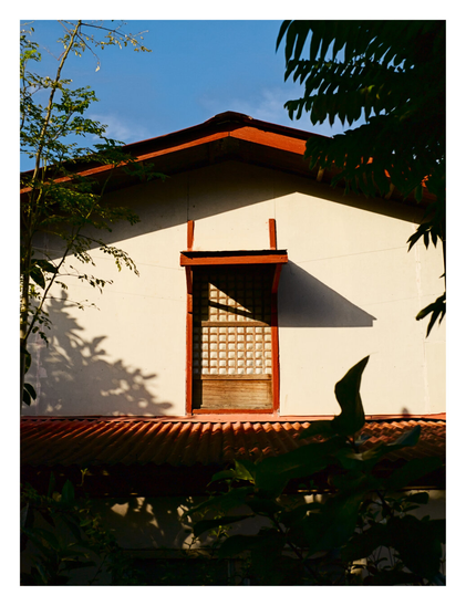 A vertical view of the upper section of a house, likely featuring traditional Filipino architectural elements. The facade is painted a creamy white and is dominated by a central window with a distinctive grid lattice pattern (resembling capiz shell panes) and a reddish-brown wooden frame.

Above and below the white wall are reddish-brown roof sections; the lower one appears to be corrugated metal. Strong sunlight casts sharp, geometric shadows from the roof eaves and the window awning across the wall. The scene is framed by green foliage on the left and right, with dark leaves in the foreground, set against a bright blue sky. - Google Gemini 3 Pro Preview