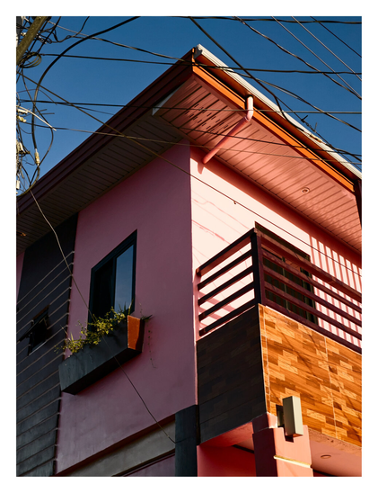 The upper corner of a vibrant pink apartment set against a deep blue sky. A chaotic web of black power lines crisscrosses the top left of the frame. The building features a balcony with dark brown railings and wood-effect siding, alongside a shadowed wall with a window box containing trailing plants. Harsh sunlight casts sharp, geometric shadows across the pink facade. - Google Gemini 3 Pro Preview