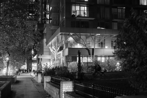 Night. Filling the upper right corner of the image is an ultramodern condo building, bright lights across its facade. At far right are shaded trees. The perspective recedes toward the left, ending in a distant patch of light that is Wellesley Station and the tiny silhouetted figures on the sidewalk outside it.