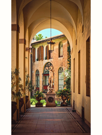 A photography of a sunlit, arched courtyard with a classical statue of a nude male figure in the center, standing on a stone pedestal. The yellow-washed walls have arched windows with wooden shutters. There are potted plants which line the pathway. A black lantern hangs from the ceiling. The background has a blue sky and greenery. The floor is tiled in earthy tones.