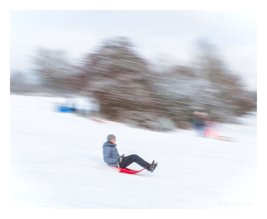 Colour photograph of a grown man tobogganing downhill on a small red plastic sled. The background scenery and figures are blurred due to the use of a slow shutter panning technique.