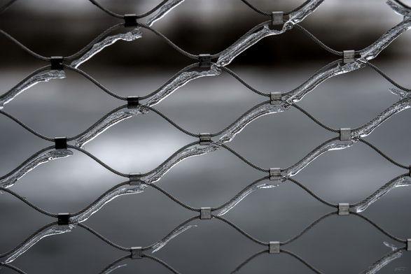 Das Foto zeigt ein vereistes Maschennetz aus Stahl. Es wirkt sowohl robust und industriell als auch ästhetisch und beinahe künstlerisch. Das Eis verleiht dem Stahldraht eine glänzende, kristalline Oberfläche, die das Licht auf interessante Weise reflektiert. So entsteht ein faszinierender Kontrast zwischen der Härte des Metalls und der zerbrechlichen Schönheit des Eises.
 ----- 
The photo shows an icy steel mesh net. It appears both robust and industrial as well as aesthetic and almost artistic. The ice gives the steel wire a shiny, crystalline surface that reflects light in an interesting way. This creates a fascinating contrast between the hardness of the metal and the fragile beauty of the ice.