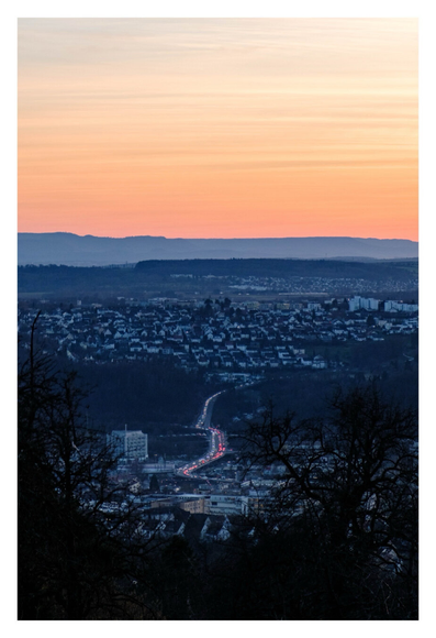Foto im Hochformat. Kurz nach Sonnenuntergang. Blick von erhöhter Position auf eine große Stadt. Im Vordergrund sind links und rechts kahle Bäume, dazwischen ist in der Ferne eine große Straße, auf der Autos Stoßstange an Stoßstange fahren. Die Straße verläuft Richtung Horizont, dort sind die Silhouetten von Bergen. Darüber ist der orangefarbene Himmel.