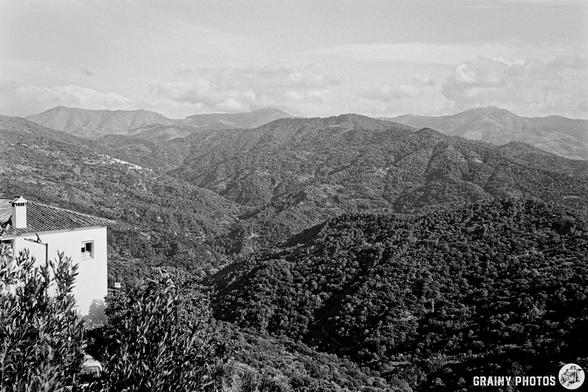 A panoramic view of rolling mountains covered in greenery, with a glimpse of a building on the left. The scene captures a serene, natural landscape under a cloudy sky. The image is presented in black and white, emphasizing textures and depth.