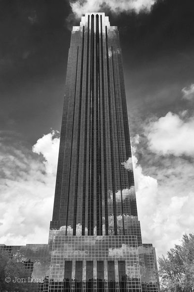 This is a black and white architectural photo in portrait format of a glass fronted high-rise office block. Houston, Texas (2012).

From the base of the image and centred in the frame, is this tall office block rising upward to just short of the top of the photo. The building has the appearance of having an almost square footprint that continues upwards to the top. The facade has an uneven but symmetrical surface with the centre having three parallel, sharp, 'V' shaped protuberances that stretch from the ground to the top. The entire facade has a highly reflective glass front that gives the building a transparent appearance with passing clouds and clear sky mirrored in the facade.

The 64 floor, 275m (902ft) Williams Tower is located in the Uptown District of Houston, Texas. 