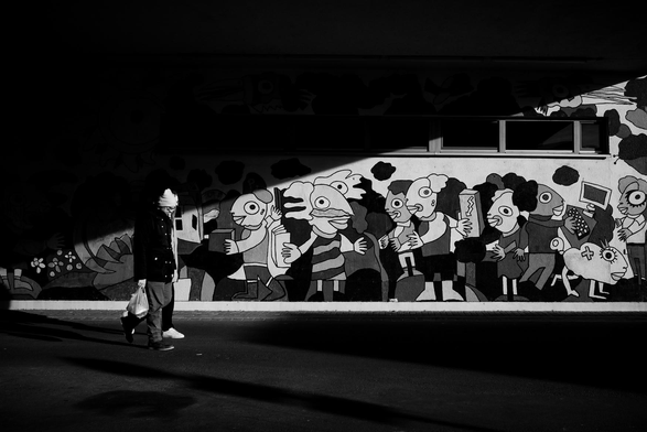 The image is a black and white photograph taken under what appears to be an overpass or a covered walkway. The scene is dominated by a brightly painted mural on the wall, partially illuminated by the sunlight streaming in. On the left side of the image, a person is walking, entering the frame from a shadowy area.

The mural depicts a series of cartoonish figures with exaggerated features. They have large eyes, often with a single pupil, and their bodies are simplified and stylized. The figures are engaged in various activities, some holding objects like a package, a box, and what seems to be a computer with a keyboard. The style is whimsical and playful, with bold outlines and flat areas of color. The mural’s overall aesthetic seems to contrast with the more mundane surroundings.