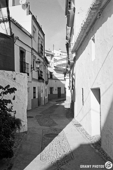 A narrow, sunlit alleyway in Algatocín, featuring white-washed buildings, textured cobblestone paths, and shadow play along the walls. The scene captures a peaceful ambiance with a blend of architectural styles.