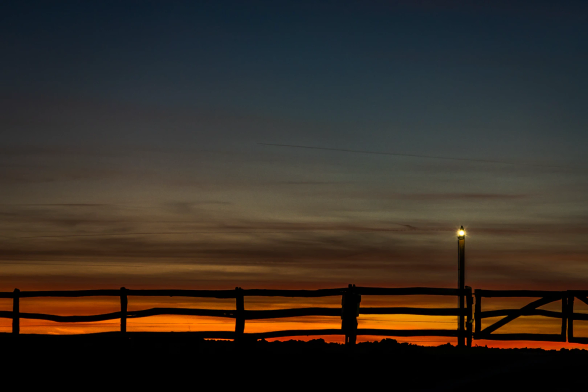 Querformat, Vordergrund sehr dunkel. Gegen den gestreiften Sonnenuntergangshimmel ein Ranchzaun als Silhouette, auf einem verlängerten Pfeiler eine halbhelle Lampe, die sich sehr bemüht.