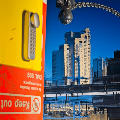 Abstract water reflection photograph showing distorted images of buildings and a yellow emergency phone box reflected in rippling water at Salford Quays, creating impressionistic patterns in blue, yellow, and orange.