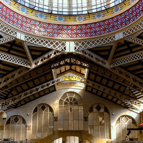 View on a detail of the interior of the central market of Valencia, Spain. At the top of the frame part of a dome with glass panes is visible. The ceiling is supported by many decorative iron beams and features beautiful red and blue tile work. The lower half of the image show a number of lovely large windows.