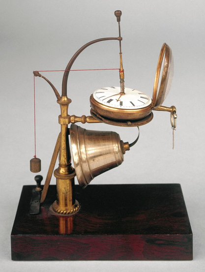 An upright turned brass column, mounted on a rectangular mahogany base, supports a horizontal carrier for a pocket watch under which is mounted a bell on a spring. The bell is raised on a lever which is held in place by a spring fixed to the base. From the centre arbor of the watch a cord has a weight on the end. The operation is similar to that of a mouse-trap - when the weight touches the spring the bell is released.