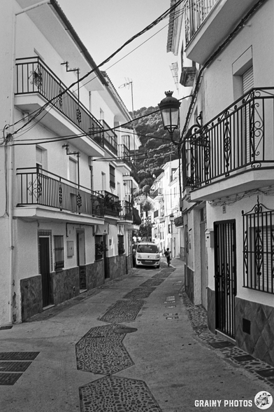 A narrow, cobblestone street in Algatocín features white buildings with balconies and ornate railings. A van is parked nearby, and steep hills are visible in the background, suggesting a picturesque and peaceful atmosphere.