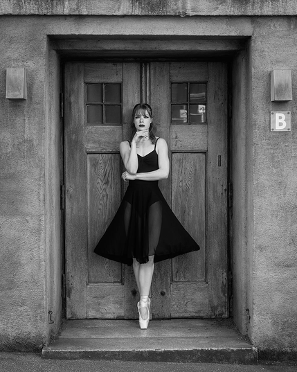 Eine junge Frau steht auf Spitzenschuhen vor einer Holztür. Sie trägt ein ärmelloses, knielanges Kleid mit transparenten Einsätzen und hält eine Hand vor den Oberkörper. Die Szene ist in schwarz-weiß gehalten. Links und rechts der Tür befinden sich rechteckige Wandlampen, rechts oben ist ein Schild mit dem Buchstaben 'B' angebracht.