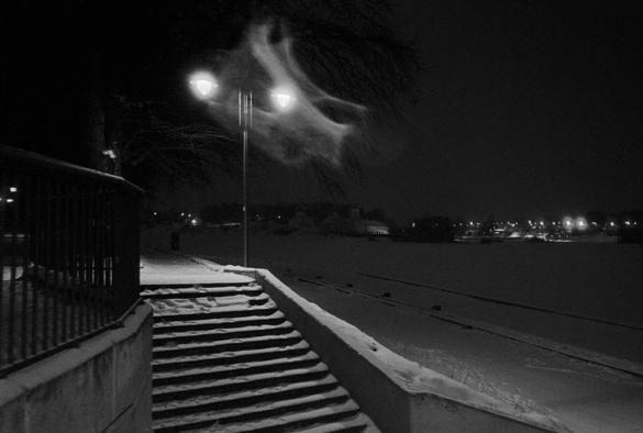 The black-and-white photo shows the embankment of the Velikaya River. Late evening. Snow is falling. A double lantern is visible in the foreground. A bright whirlwind appears in the rays of the lantern, looking like a ghost.