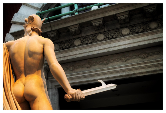 A view of the back of the marble statue “Perseus with the Head of Medusa” (1804-6) by Antonio Canova, at the Metropolitan Museum of Art in New York. The statue stands off-center to the left, seen from the buttocks up. Perseus is naked except for a flowing drape over his left arm, which extends out of frame. He wears a winged helmet, his curly hair peeking out as he looks left. His right arm hangs low, the hand gripping a short sword with a hook protruding from the blade’s edge. In the background is a shadowy doorway surrounded by an enormous stone frame with carved ornamentation. Above is a glimpse of the curve of a stone arch and some green metal bars. Sunlight from an unseen skylight above falls on the statue, accentuating its musculature and giving it a warm-toned glow.