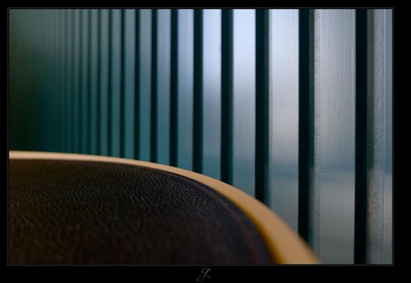 
The image shows part of the surface of a black round table with a gold metal rim in front of a petrol-colored wooden wall. The view is directed toward the wooden wall in the background, from slightly above the table surface. The table covers the lower left part of the picture, it has a rough, leather-like texture. The wooden wall consists of evenly spaced vertical slats on a wooden surface, forming a pattern of petrol-colored planes with dark shadows in the spaces between them. Indirect sunlight is gently reflected on the wooden surface, creating a smooth transition from light to dark towards the left edge of the image. Only a thin focal plane extends horizontally across the lower third.

AI disclaimer: Using my work, its meta data, written or derived description to create media with or train AI based systems is prohibited.