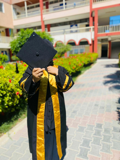 Naeema in graduation attire, obscured face holding black mortarboard, standing on a brick walkway with green shrubbery and a building in the background.