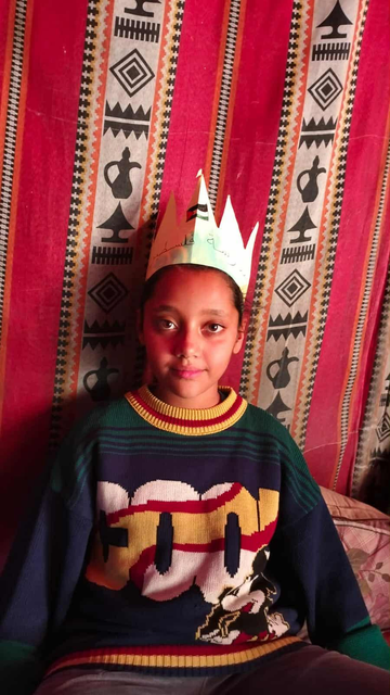 Young girl (Aylan) with dark hair and skin wearing a paper crown with Arabic writing, and a colorful patterned sweater, posing in front of a red, patterned textile backdrop.