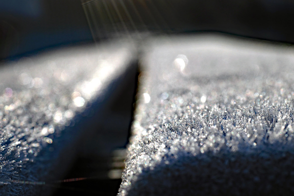 Nahaufnahme von Frost auf einer Parkbank: Die Sonne lässt die unzähligen Eiskristalle auf den dunklen Holzlatten funkeln. Ein paar Lichtstrahlen fallen von oben auf die Sitzfläche. Der Fokus liegt auf dem vorderen Drittel, der Rest des Bildes ist eher unscharf. Das Bild wirkt monochrom schwarz-weiß, ist aber tatsächlich eine Farbfotografie.

Close-up of frost on a park bench: the sun makes the countless ice crystals on the dark wooden slats sparkle. A few rays of light fall from above onto the seat. The focus is on the front third, the rest of the image is rather blurred. The image appears monochrome black and white, but is actually a colour photograph.