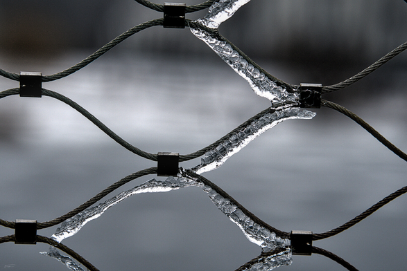 Das Foto zeigt ein Netz aus geflochtenen Stahlkabeln, die sich kreuz und quer überspannen. An den Kreuzungspunkten sind Metallklammern befestigt, die die Kabel zusammenhalten. Einige der Kabel sind mit einer glänzenden Eisschicht überzogen. Der neutrale, graue Hintergrund hebt die Struktur der Kabel besonders hervor. Insgesamt wirkt die Aufnahme technisch und industriell.
 ------- 
The photo shows a network of braided steel cables crisscrossing each other. Metal clamps are attached at the intersections to hold the cables together. Some of the cables are covered with a shiny layer of ice. The neutral gray background particularly highlights the structure of the cables. Overall, the photo has a technical and industrial feel.