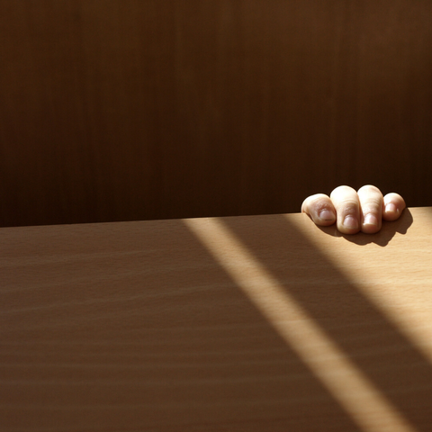 A photo divided in two: a section of wooden chair at the top and part of a lighter wooden table at the bottom. At the right four small fingers grip the tabletop, bathed in a strip of sunlight.