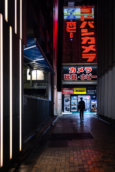 A dark little pedestrian street at night in Shinjuku in Tokyo bathed in a cyberpunk atmosphere. The only sources of light come from gigantic red neon letters in japanese characters set vertically above a backside entrance of the famous Yodobashi camera store in the center of the image, and other white neon bars on a dark building on the left of the image . A man dressed in black is walking in the dark towards the lit entrance, his little silhouette contrasting with the light source.