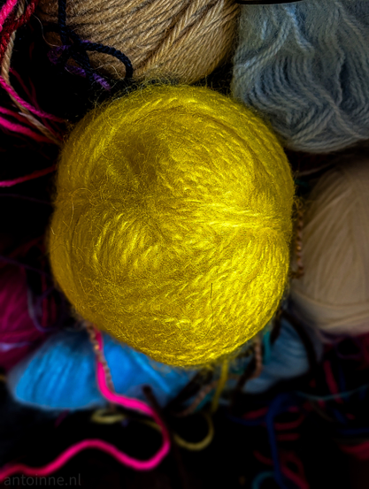A vibrant, close-up photograph featuring a collection of yarn skeins, with a focus on color and texture.

The central focus is a large, bright yellow ball of yarn. It has a distinctly fuzzy, mohair-like texture with many fine fibers catching the light, giving it a soft, almost glowing appearance. 

The ball is positioned slightly above the center of the frame. The yellow yarn is surrounded by several other skeins in various muted and bold tones.